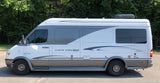 Side view of a modern white and gray Vista Cruiser camper van, an RV rental, parked on asphalt with trees in the background.