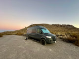 Beautiful sunset over an rv rental: a dark grey Sprinter van parked on a dirt road in a desert landscape with hills and moon.