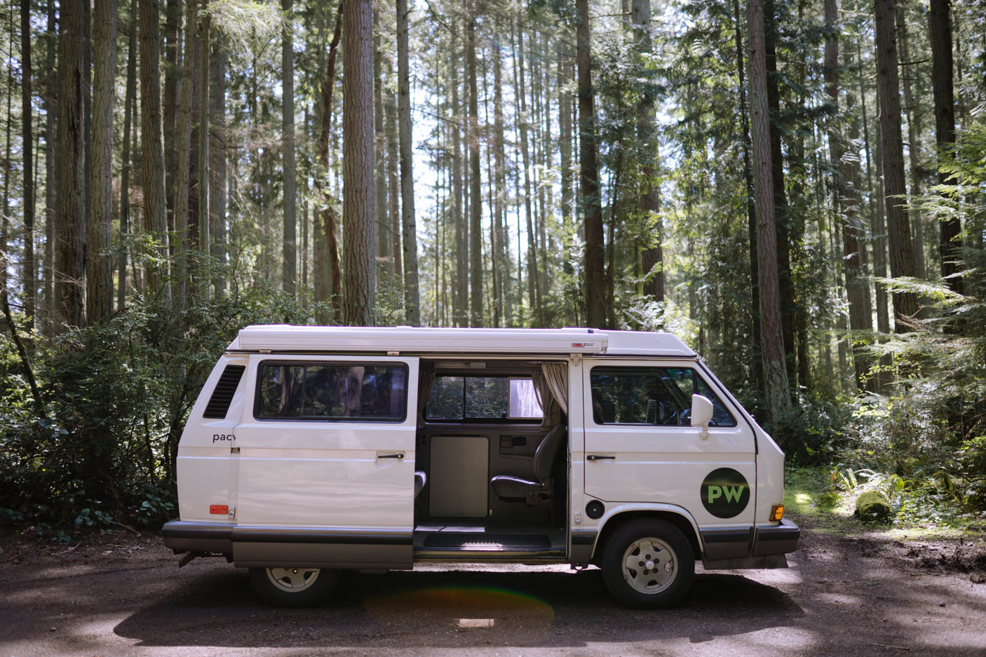 A white camper van, ideal for RV rental adventures, parked in a sunny forest with its side door open.