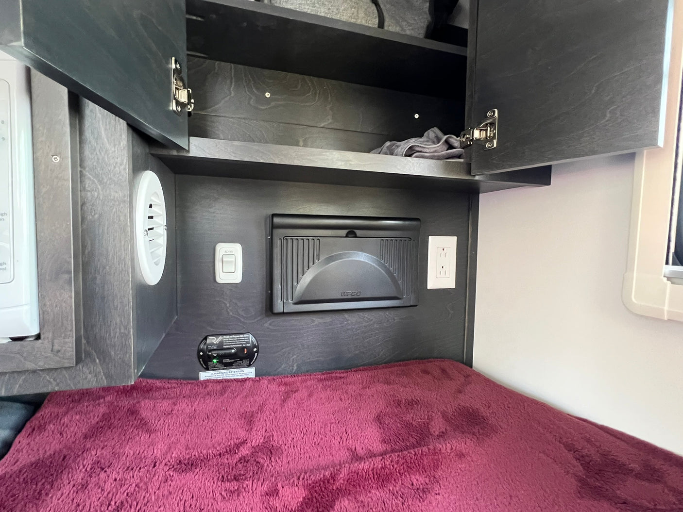 RV rental bedroom view: Dark wood-grain cabinets open above a bed. A microwave, power panel, and safety alarm are installed.
