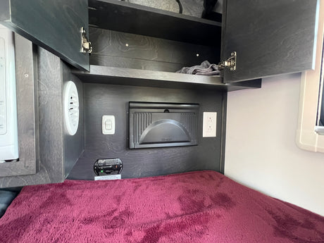 RV rental bedroom view: Dark wood-grain cabinets open above a bed. A microwave, power panel, and safety alarm are installed.