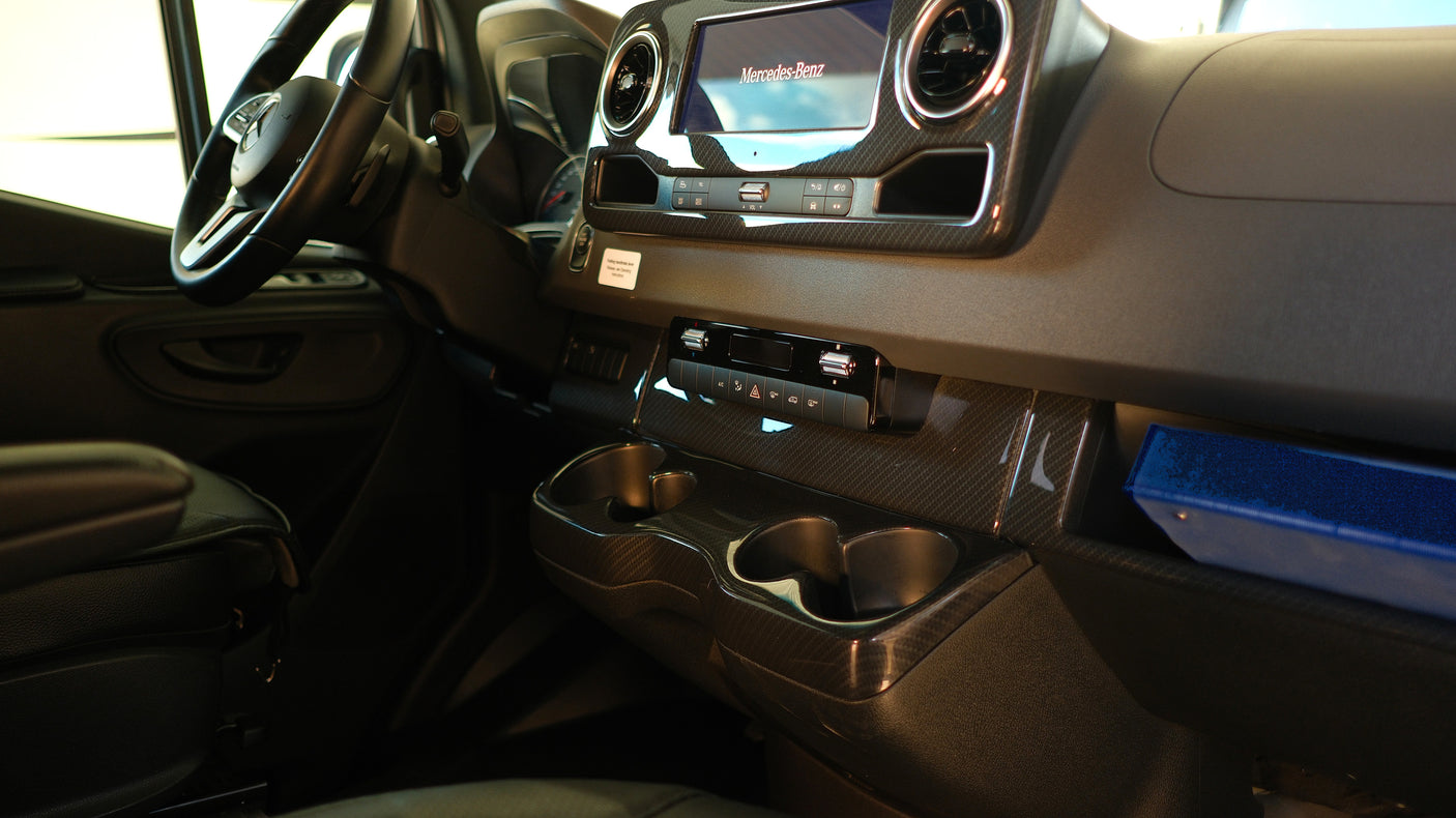 Inside a modern Mercedes-Benz rv rental: dashboard, steering wheel, infotainment screen, carbon fiber-look trim, cup holders, and storage.