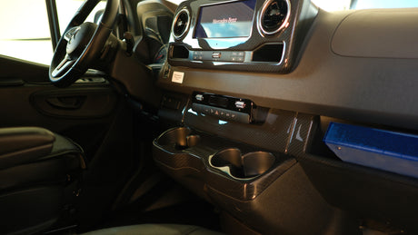 Inside a modern Mercedes-Benz rv rental: dashboard, steering wheel, infotainment screen, carbon fiber-look trim, cup holders, and storage.