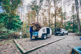 Cozy RV rental campsite in the forest: A person cooks at a teardrop trailer, with a truck & string lights nearby.