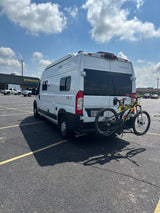 White RV rental with a yellow mountain bike on its rear, parked in a sunny asphalt lot with striped parking spaces.