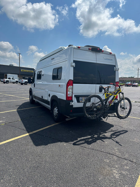 White RV rental with a yellow mountain bike on its rear, parked in a sunny asphalt lot with striped parking spaces.