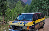 This colorful RV rental van is parked on a dirt road in a lush mountain forest, ideal for camping adventures. A small dog is nearby.