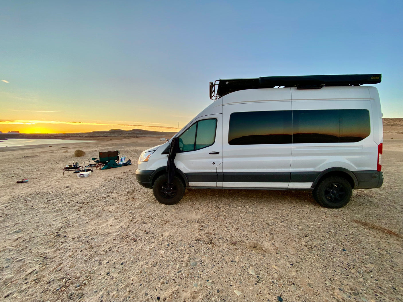 White camper van (RV rental) enjoying a desert lake sunset with a cozy camp setup. Ideal for off-grid adventure.