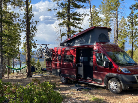 Scenic campsite with a red RV rental, two mountain bikes, and a hammock among pines, overlooking a valley.