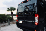 Rear of a black Thor Sequence RV rental van with a bike rack, parked by water and a palm tree, at dusk.