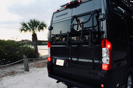 Rear of a black Thor Sequence RV rental van with a bike rack, parked by water and a palm tree, at dusk.
