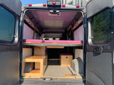 RV rental: Interior of a camper van with open back doors, showcasing a raised bed with purple cushions and wood storage.