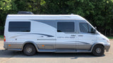 Side view of a white and grey camper van RV rental, a Vista Cruiser MB Edition, parked outdoors among trees.