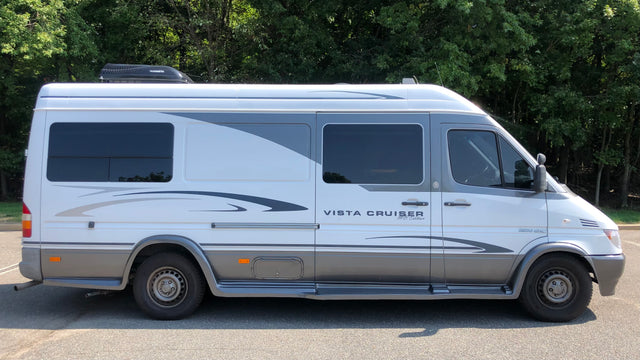 Side view of a white and grey camper van RV rental, a Vista Cruiser MB Edition, parked outdoors among trees.