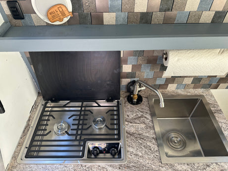Compact RV rental kitchenette features a two-burner stove, stainless steel sink, and paper towel holder, ideal for camper cooking.