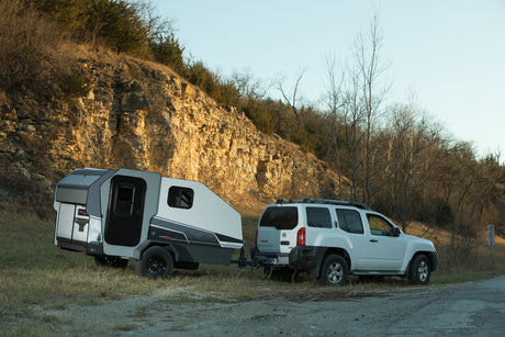 White Nissan Xterra SUV towing a grey and black camper trailer (rv rental) by a rocky cliff, ideal for outdoor travel.