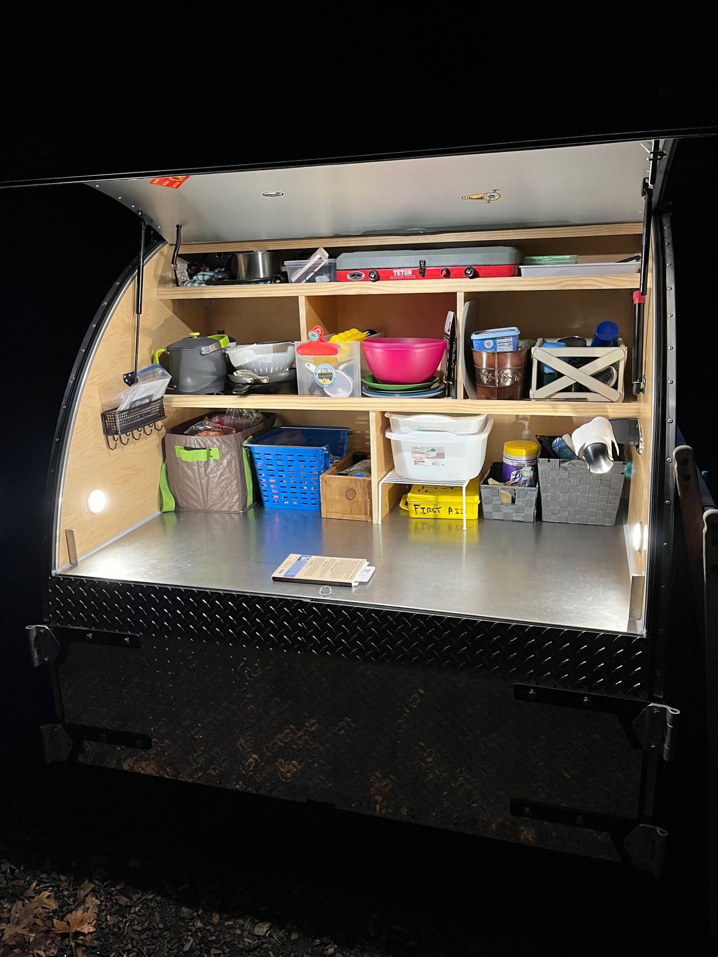 Night shot of an organized teardrop RV rental kitchen, ready for camping. Shelves hold cooking gear & supplies.