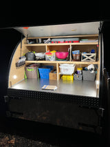 Night shot of an organized teardrop RV rental kitchen, ready for camping. Shelves hold cooking gear & supplies.