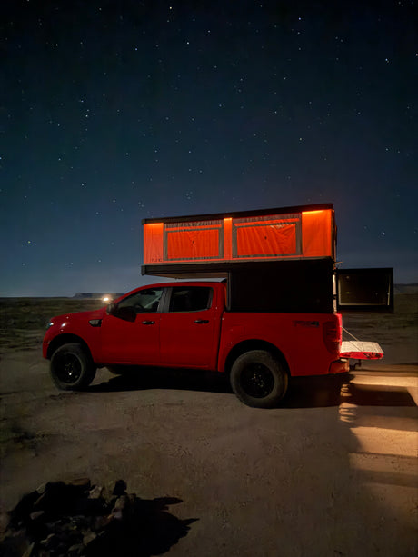 An RV rental red pickup truck with an illuminated rooftop camper under a clear, starry night sky. Outdoor adventure awaits.
