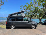 A dark grey camper van RV rental, with its pop-top open, is parked by the turquoise ocean for scenic views.