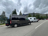 Dark grey and white RV rental vans parked by a scenic mountain lake under cloudy skies.