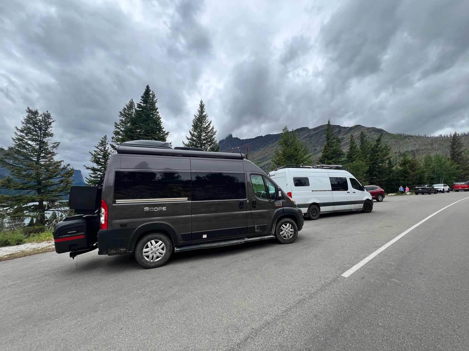 Dark grey and white RV rental vans parked by a scenic mountain lake under cloudy skies.
