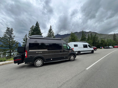 Dark grey and white RV rental vans parked by a scenic mountain lake under cloudy skies.