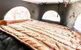 Inside an RV rental: a snug bed with a textured brown blanket, offering bright views from arched pop-top windows.