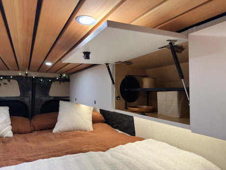Cozy RV rental interior featuring a wooden ceiling, comfortable bed, and open overhead storage cabinets. Ideal for a campervan trip.
