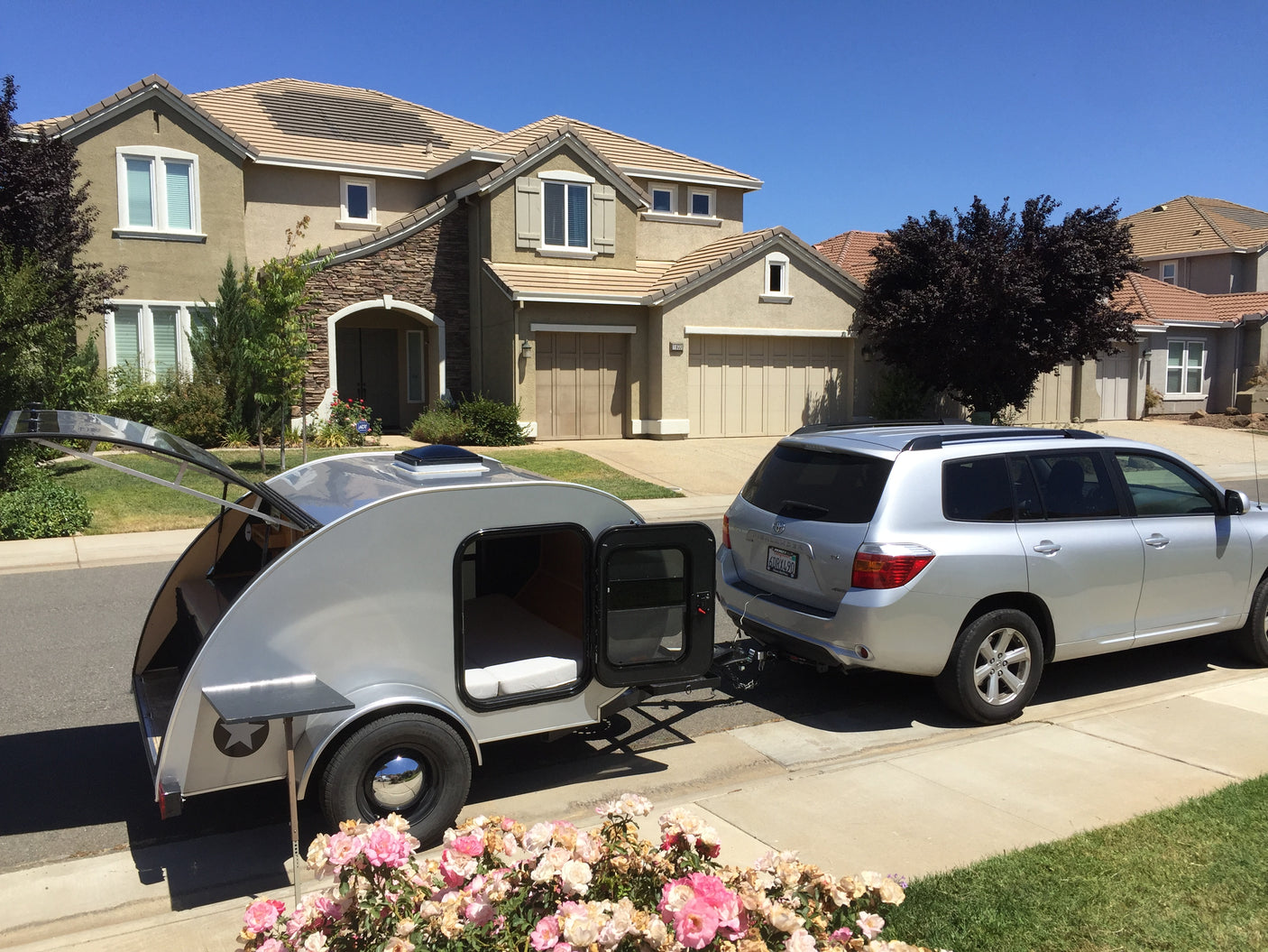A silver teardrop camper (rv rental) is hitched to an SUV on a sunny residential street in front of a house.