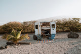 Enjoying an RV rental setup. A man relaxes in a white camper van with a surfboard, chairs, and mat on a gravel area. Palm tree close.