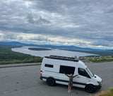 White Sprinter RV rental at scenic overlook. Happy woman enjoys vast lake, forest, & mountain views under cloudy skies.