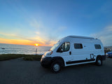 White Winnebago Solis RV rental parked oceanfront at sunset. Golden sun dips below horizon, waves crash on shore. Ideal for travel.