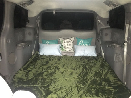 Cozy vehicle bed setup in a gray minivan/SUV, with a green sleeping bag and decorative pillows. Great for car camping or an RV rental experience.