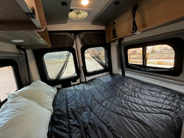 Cozy RV rental interior: bed, windows with nature views, overhead storage. Perfect for road trips and camping adventures.