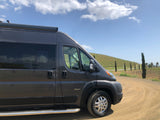 A dark gray Jayco RV rental camper van on a dirt road, next to a sunny vineyard with cypress trees. Perfect for a rural trip.