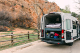 RV rental camper van's rear opens to a cozy bed and storage, parked roadside next to a rustic fence with vibrant red rock cliffs in the background.