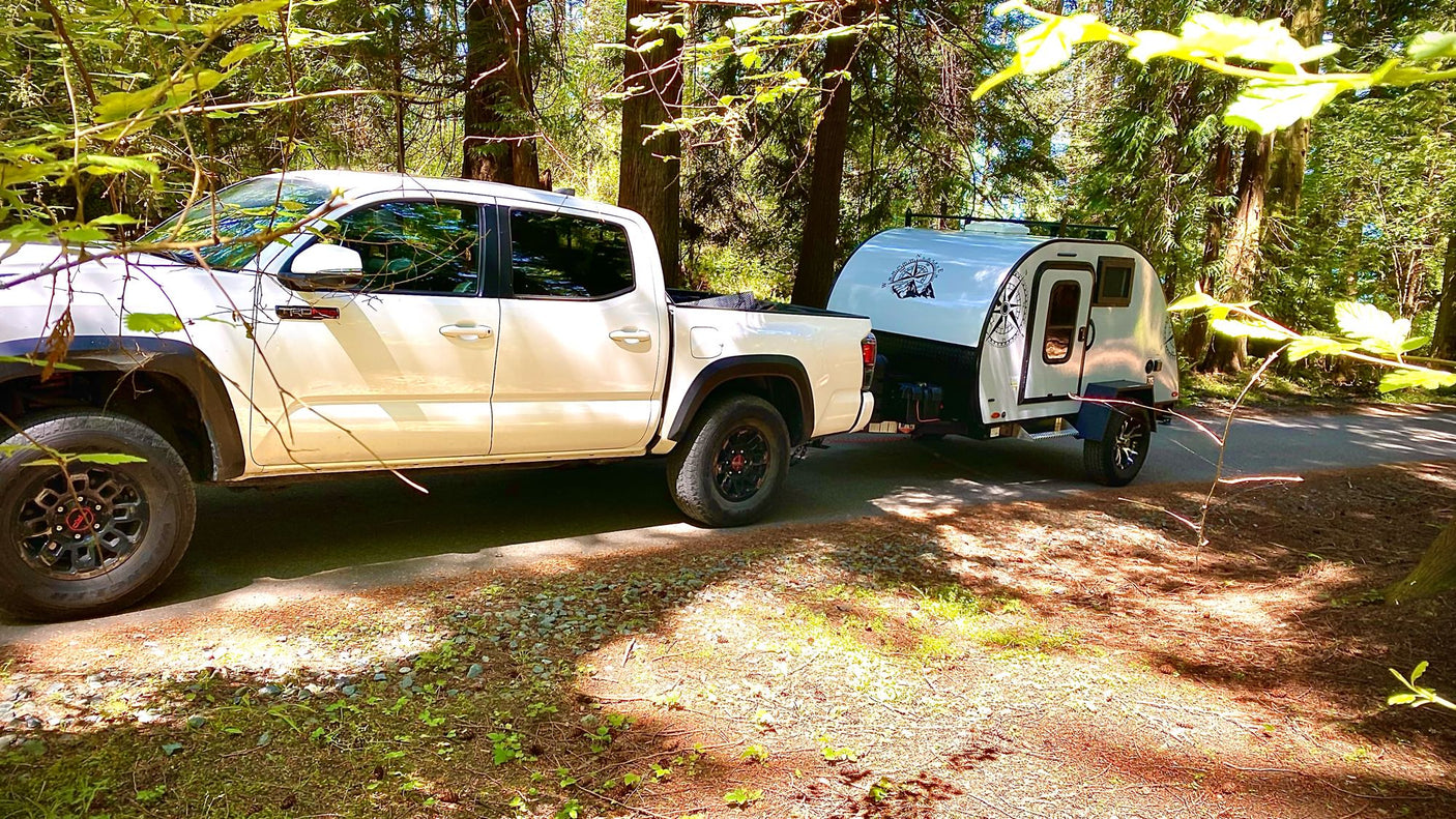 White truck towing a small teardrop camper in a sunny forest. Ideal RV rental for your next outdoor adventure.