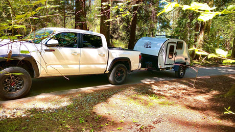 White truck towing a small teardrop camper in a sunny forest. Ideal RV rental for your next outdoor adventure.