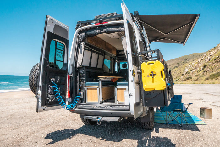 Rear view of an RV rental camper van at the beach, doors open revealing interior, water/fuel tank, and awning.