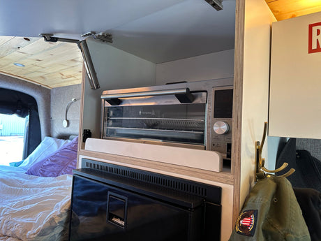 RV rental kitchen area with a built-in toaster oven, an Isotherm fridge, and a bed. Cabinet has a lift-up door.