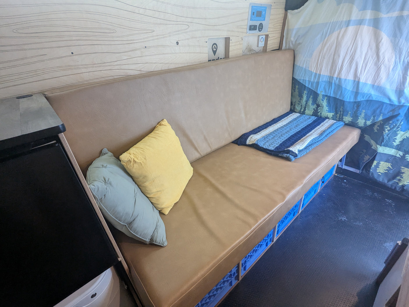 RV rental interior: Light brown bench, pillows, striped blanket, blue crate storage, wood wall, and control panel.