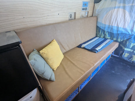 RV rental interior: Light brown bench, pillows, striped blanket, blue crate storage, wood wall, and control panel.