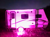 A white RV rental glows vibrantly with pink light at night, parked in a dark, grassy outdoor scene.