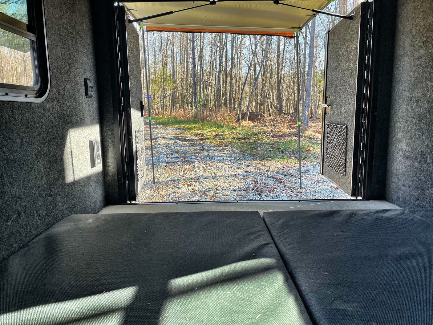 Cozy rv rental interior: dark beds face an open rear door, revealing a sunny gravel path leading into a bare forest.