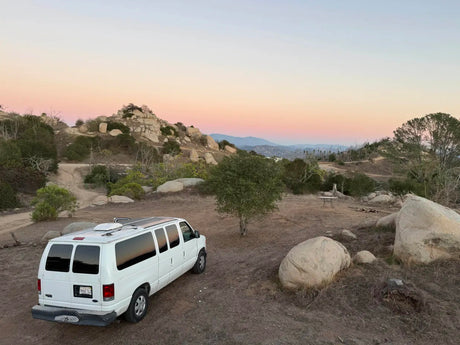 Scenic RV rental camper van parked overlooking hills and large boulders. A vibrant sunset paints the sky, ideal for outdoor adventures.