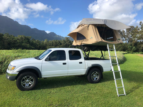 A white pickup truck with a tan rooftop tent and ladder, parked on green grass with mountains. Ideal for an RV rental adventure.