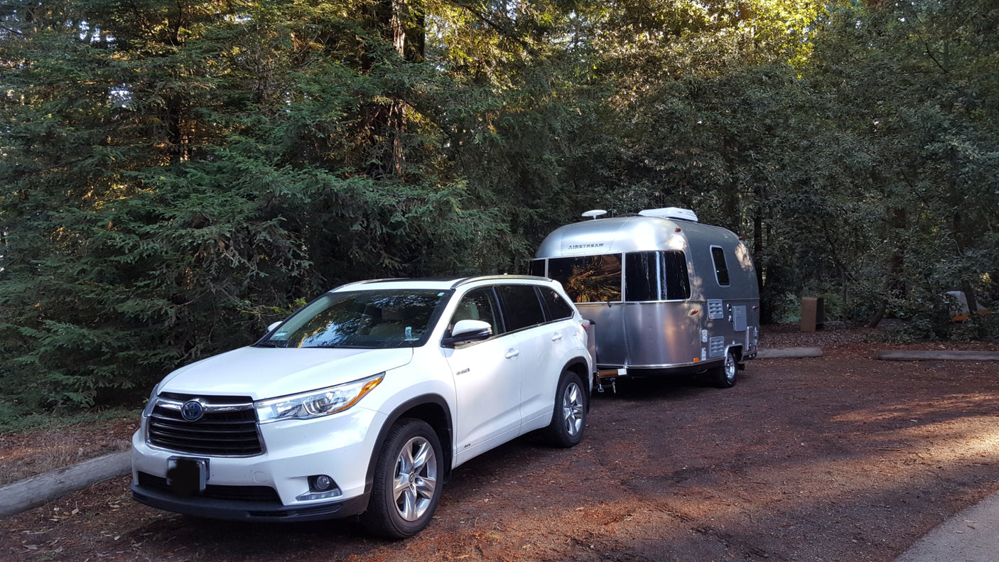 White SUV towing a silver Airstream travel trailer, likely an rv rental, parked in a shaded wooded campsite.