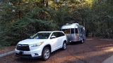 White SUV towing a silver Airstream travel trailer, likely an rv rental, parked in a shaded wooded campsite.