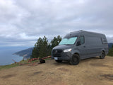Gray Mercedes-Benz Sprinter camper van (RV rental) and dog overlooking a scenic ocean coast.